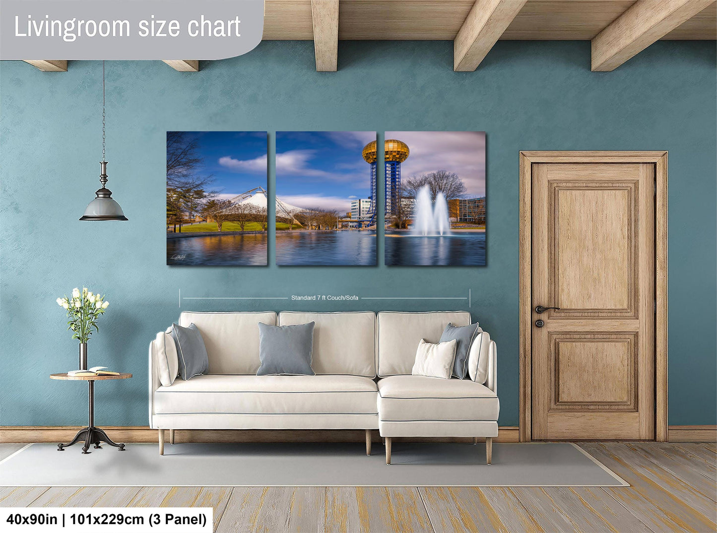 The image is a living room with a white sofa and a wooden door. Above the sofa, there is a large living room size chart that provides dimensions for the artwork displayed on the wall. The artwork is a collage of three pictures depicting a cityscape with a tall tower and a body of water.