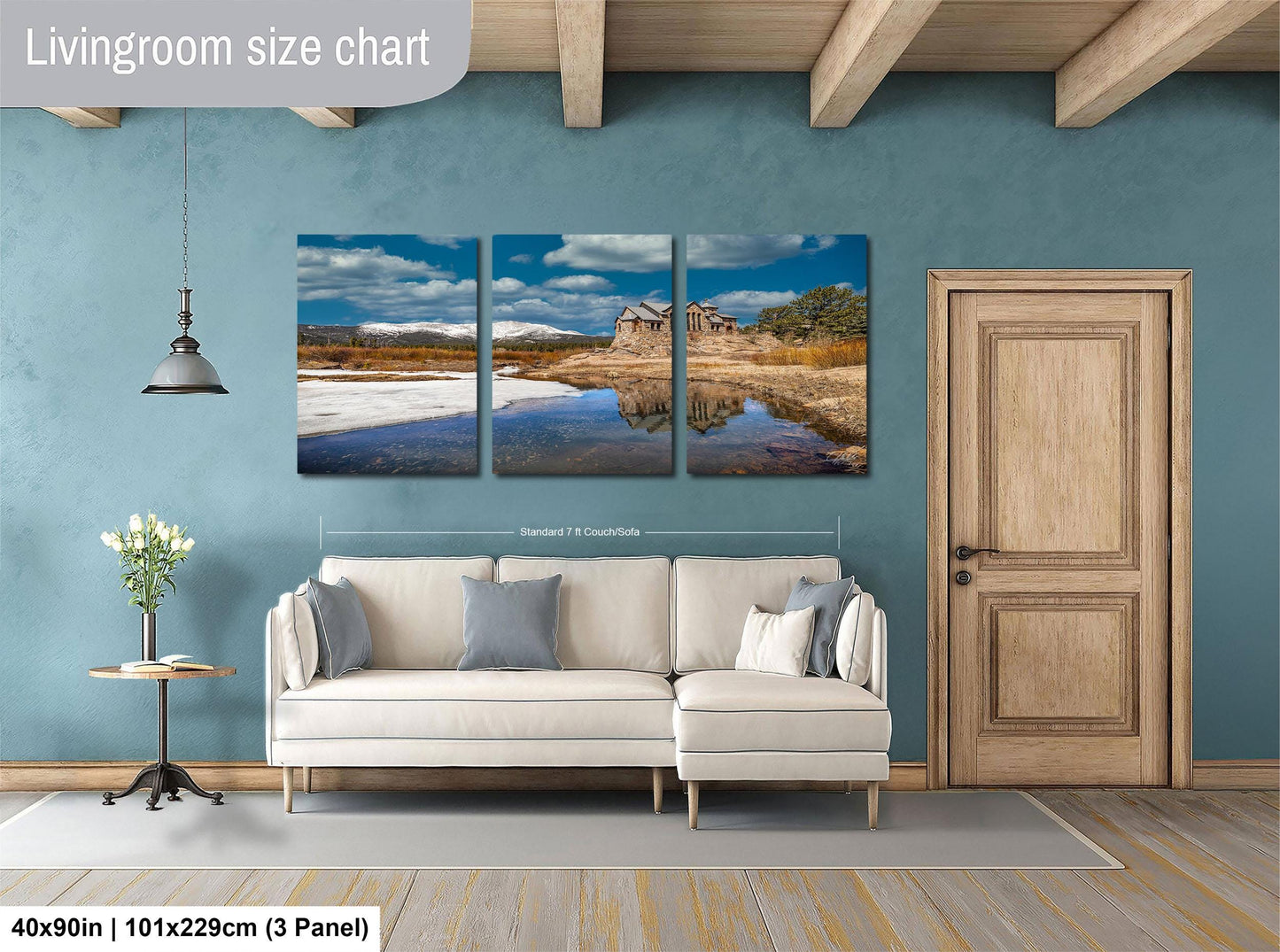 The image is a living room with a large wall art piece depicting a scenic landscape with a lake and mountains. The living room has a white sofa and a wooden door, and there is a potted plant and a vase on a side table. The wall art is approximately 40 inches wide and 101/129 centimeters tall, and it is a three-panel collage.