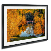 A framed photograph depicting a serene autumn landscape with a bridge reflected in a calm body of water, surrounded by trees with vibrant orange and yellow foliage.