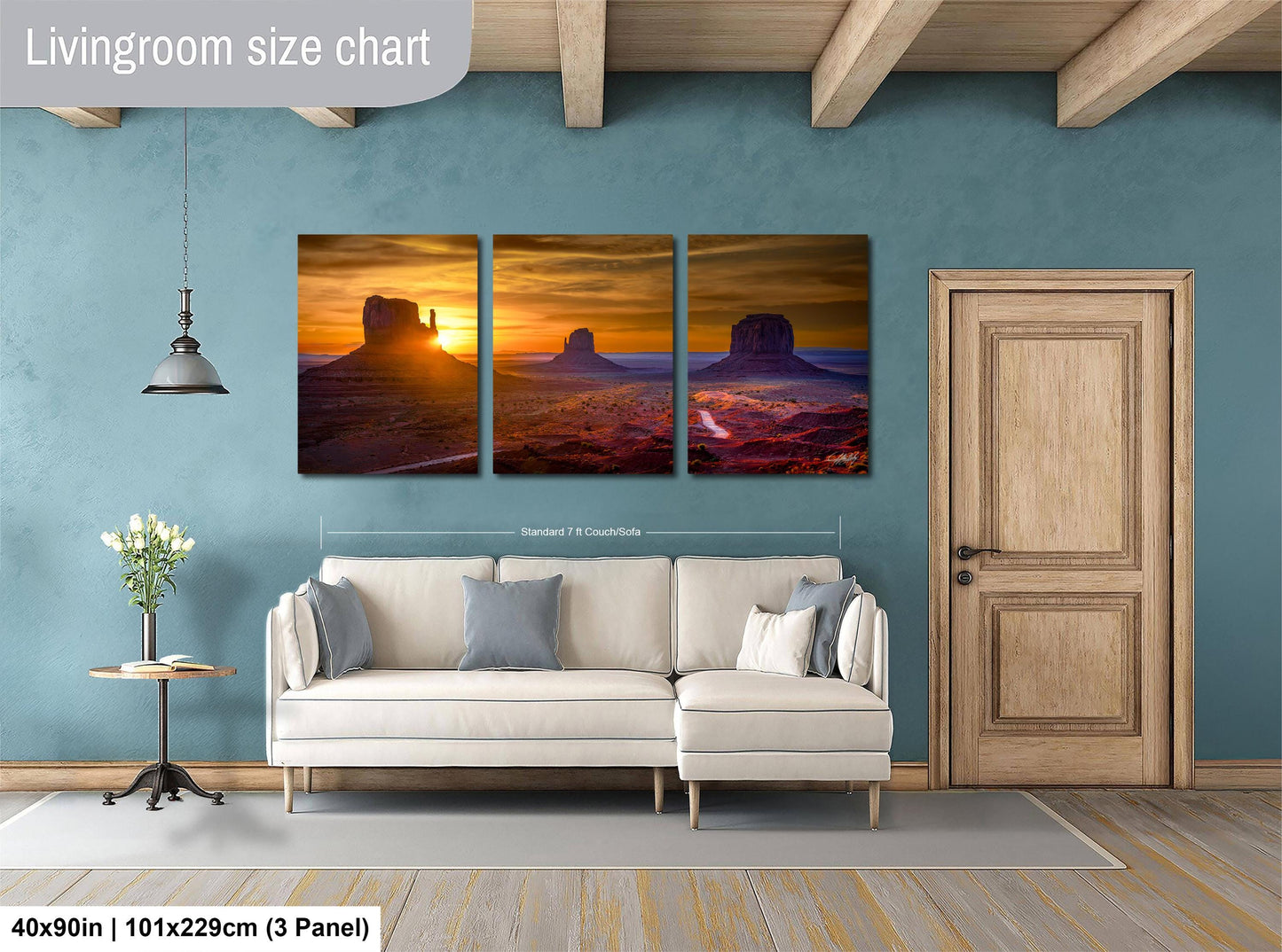 The image is a livingroom with a large wall art depicting a desert landscape with mountains and a sunset. The wall art is divided into three panels, each showing a different stage of the sunset. The living room has a white sofa and a wooden door, and there is a potted plant on a side table. The wall art is approximately 40 inches wide and 101.29 cm tall, and it is mounted on a wall with a wooden frame.
