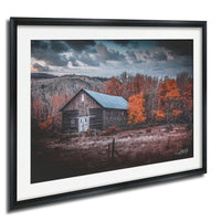 A rustic wooden barn with a sloping roof stands in a field surrounded by trees with vibrant orange leaves, set against a dramatic cloudy sky.