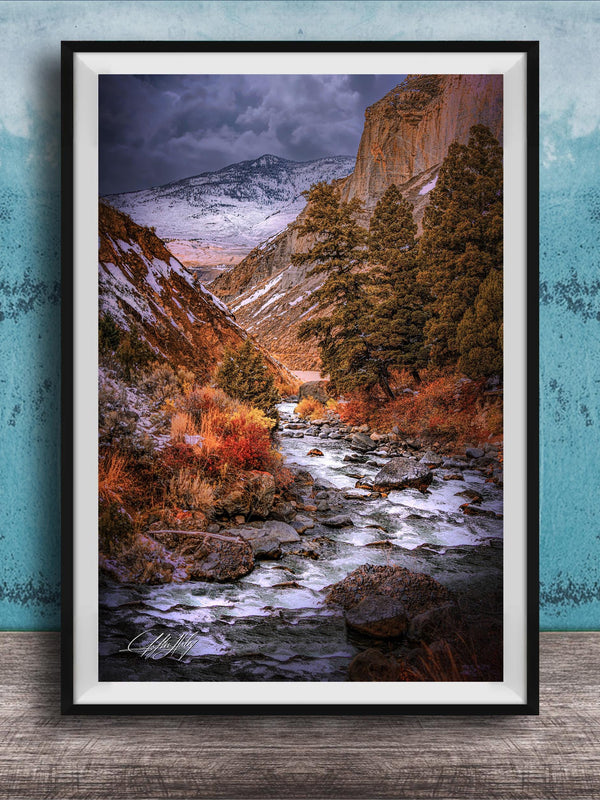 The image is a framed photograph of a scenic mountain landscape with a winding river flowing through it. The mountains are covered in snow, and the sky is filled with clouds, creating a dramatic and moody atmosphere. The photograph is framed and displayed on a wooden surface, with a blue wall in the background.