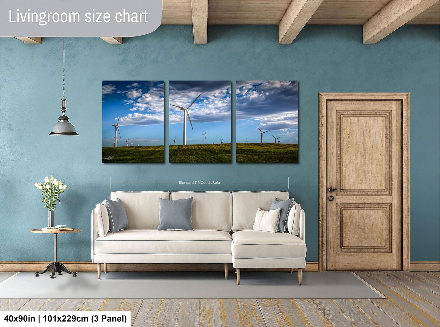 The image is a living room with a large white sofa, a wooden door, and a hanging light fixture. Above the sofa, there is a livingroom size chart that provides dimensions for the artwork displayed on the wall. The artwork consists of three panels depicting wind turbines against a blue sky with clouds.