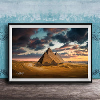 A framed photograph of the iconic pyramids of Giza against a dramatic sky at sunset, with the sun setting behind the sand dunes.
