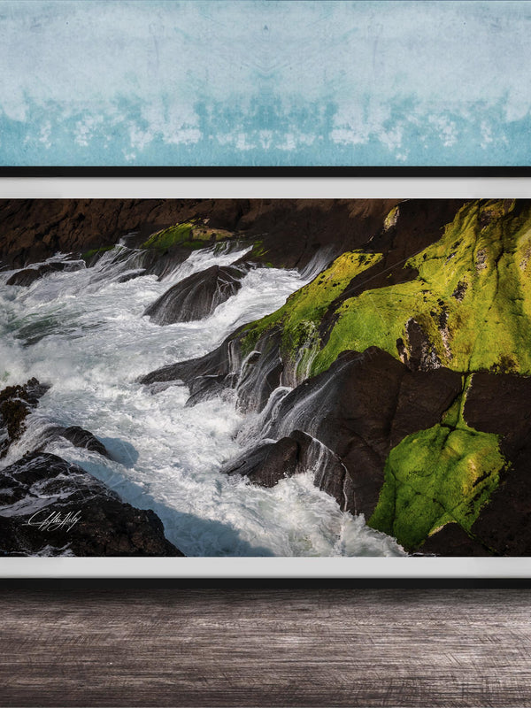 A framed photograph depicting a rocky coastline with flowing water and patches of green moss.