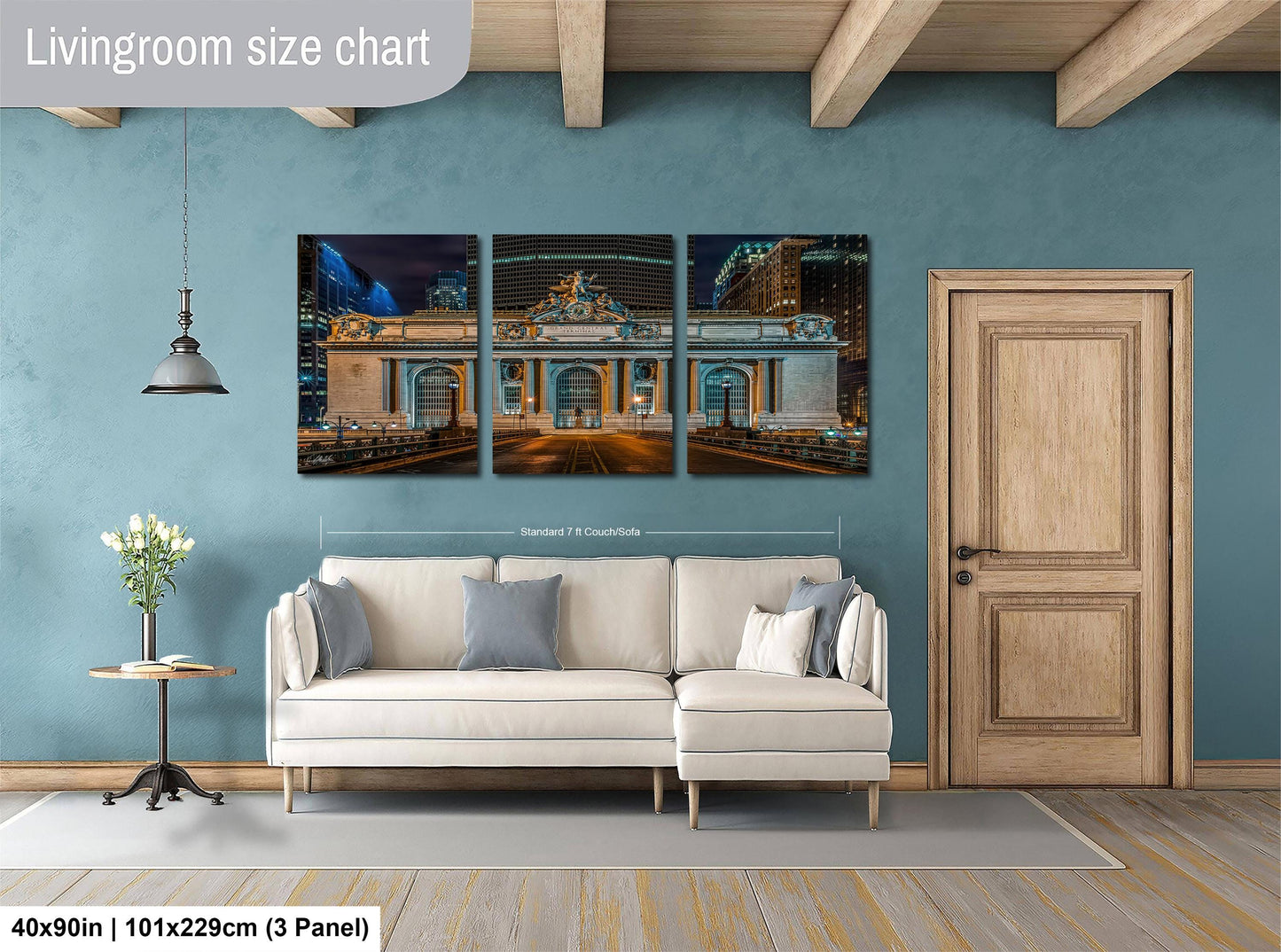 The image is a living room with a large, three-panel wall art depicting a cityscape at night. The wall art is mounted above a white sofa, and there is a wooden door and a small table with a vase of flowers nearby. The wall art is approximately 40 inches wide and 101.29 cm tall, with each panel measuring approximately 3 feet in width.