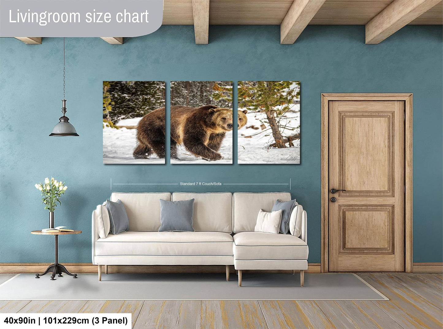 The image is a living room with a large wall art depicting a brown bear walking through a snowy forest. The wall art is divided into three panels, each showing a different angle of the bear in the snowy landscape. The living room has a white sofa and a wooden door, creating a cozy and inviting atmosphere.
