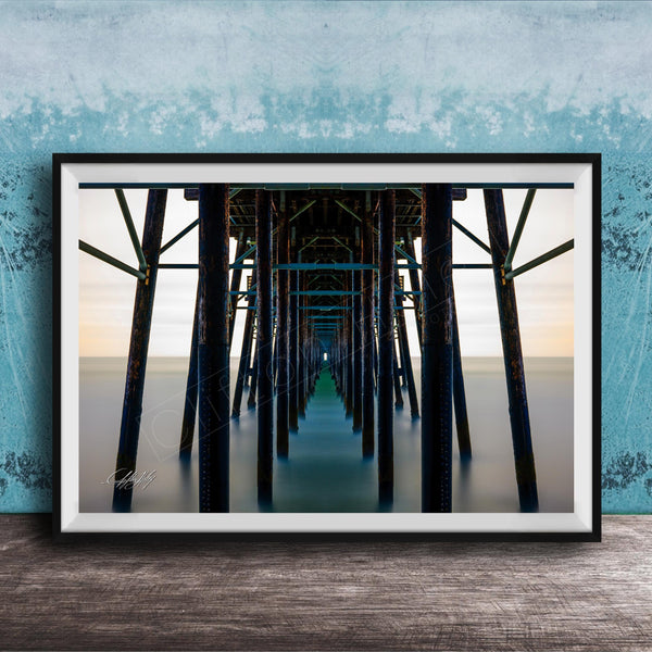 The image is a framed photograph of a long pier with multiple wooden columns extending into the distance, creating a sense of depth and perspective.