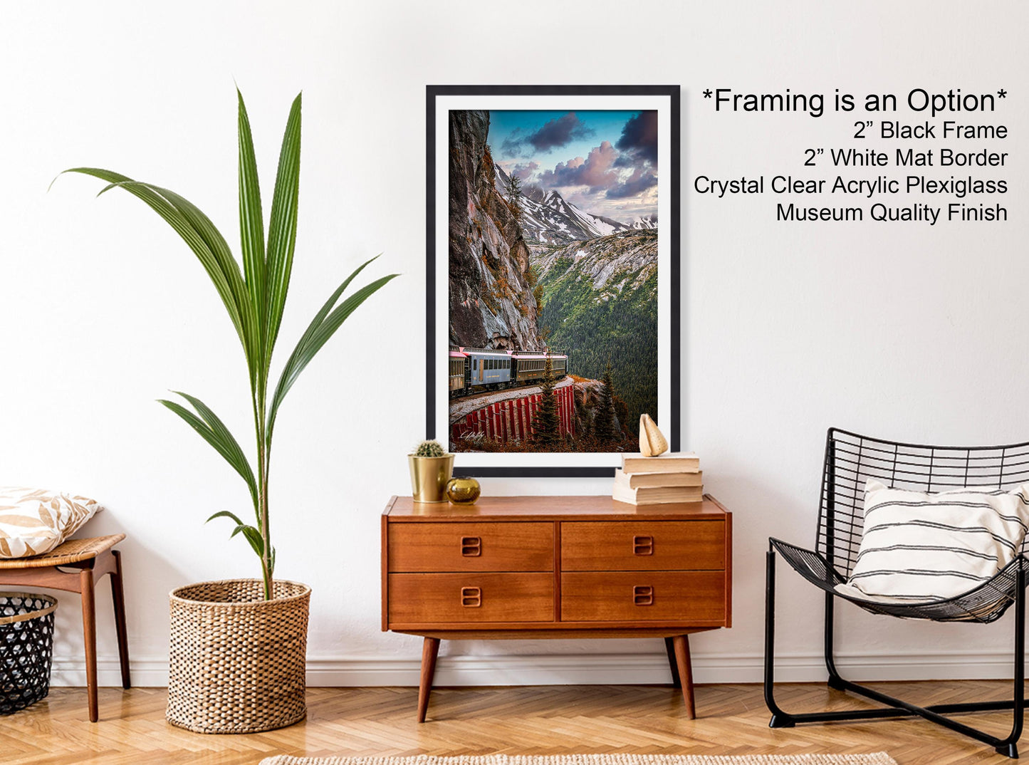 a framed photograph of a train on a mountain, hanging on a wall in a room with a wooden dresser, a woven basket, and a black chair.