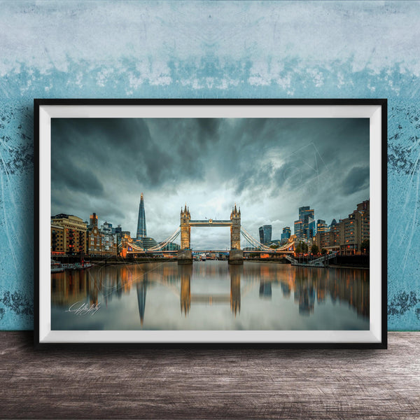 A framed photograph of the Tower Bridge in London, England, with the city's skyline reflected in the water below. The sky is filled with clouds, creating a dramatic atmosphere.