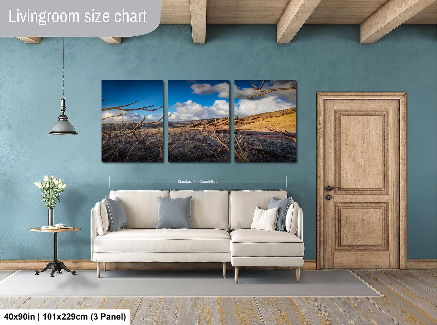 The image is a living room with a white sofa and a wooden door. Above the sofa, there is a large wall art piece depicting a desert landscape with a blue sky and clouds. The wall art is divided into three panels, each showing different aspects of the desert scene. The living room has a blue wall and a wooden floor, creating a cozy and inviting atmosphere.