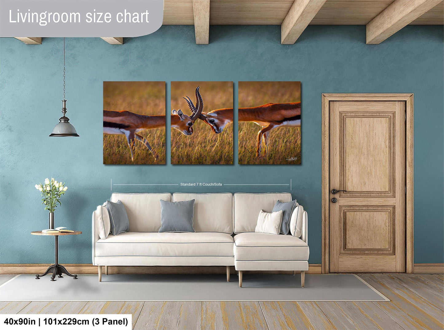 The image is a living room with a large white sofa, a wooden door, and a painting of two gazelles in a field. The painting is divided into three panels, and the wall behind the sofa is painted a light blue color.