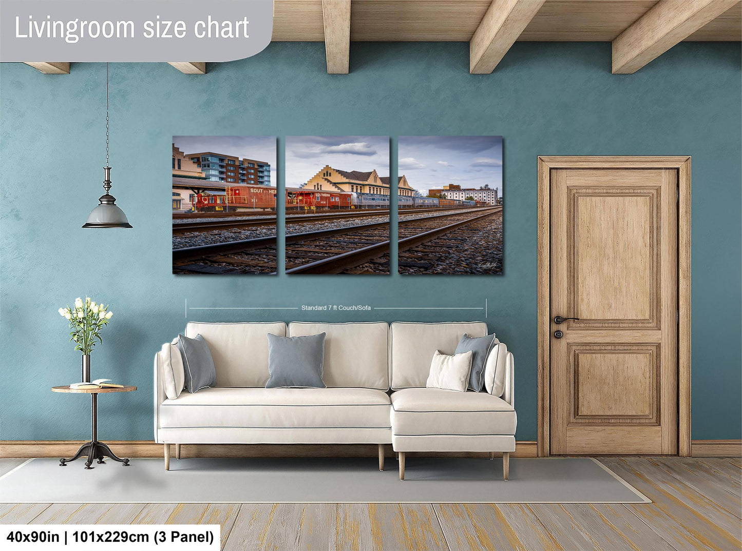 The image is a living room with a white sofa and a wooden door. Above the sofa, there is a large wall art depicting a train station with multiple train tracks. The wall art is divided into three panels, each showing different parts of the train station. The wall art is approximately 40 inches wide and 101.29 cm tall, with a 3-panel arrangement.