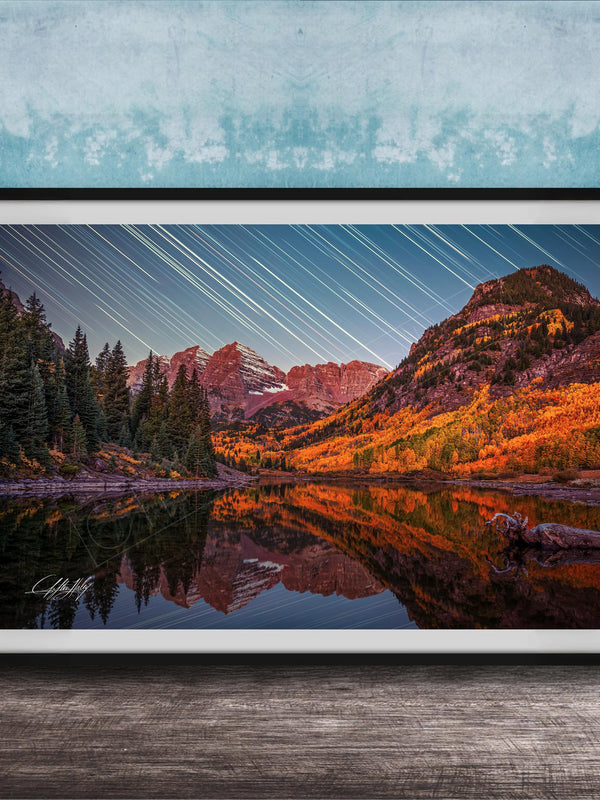 The image is a framed photograph depicting a serene mountain landscape with a lake reflecting the vibrant colors of the autumn foliage. The sky is filled with long exposure star trails, creating a stunning visual effect that adds depth and dimension to the scene.