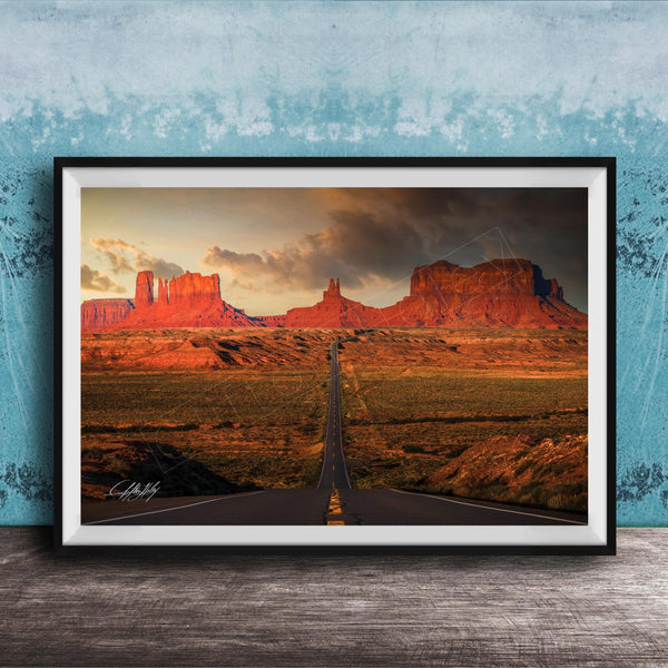 A framed photograph depicting a desert landscape with a winding road leading towards a series of red rock formations in the distance, set against a cloudy sky.