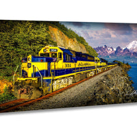 A yellow and blue Alaska train travels along a track beside a body of water, with mountains in the background.