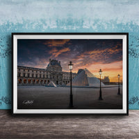 A framed photograph of the Louvre Museum in Paris, France, with the iconic glass pyramid structure of the Louvre Museum prominently featured against a dramatic sky at sunset.