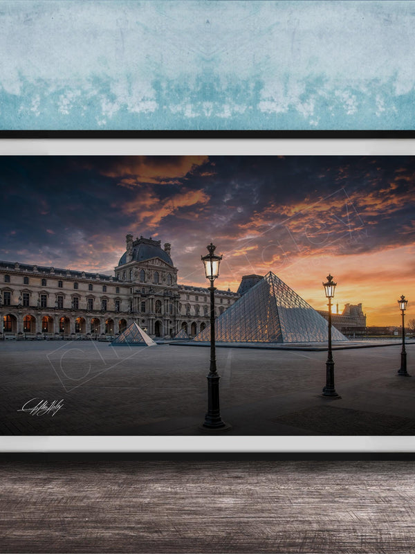 A framed photograph of the Louvre Museum in Paris, France, with the iconic glass pyramid structure of the Louvre Museum prominently featured against a dramatic sky at sunset.