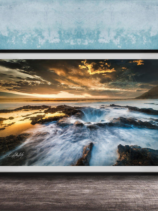 The image is a framed photograph of a rocky coastline with crashing waves, set against a dramatic sky with clouds and a sunset.