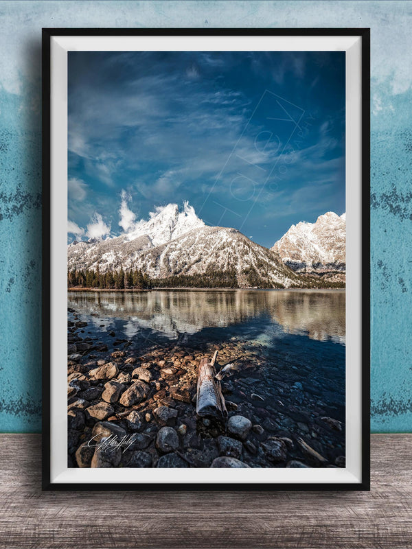 A framed photograph of a serene mountain landscape with a calm lake in the foreground, surrounded by snow-capped peaks and a clear blue sky.