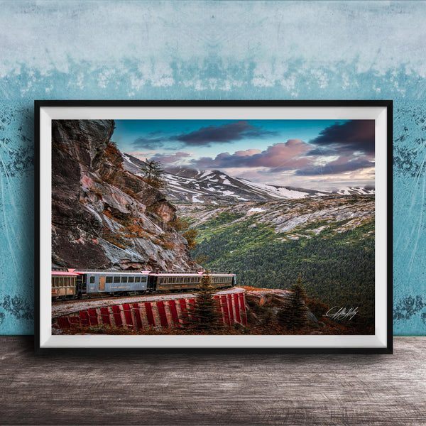 A framed photograph depicting a train traveling along a winding track through a mountainous, forested landscape with snow-capped peaks in the background.