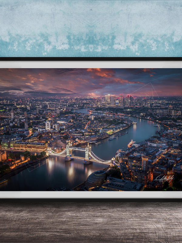 The image is a framed aerial view of a city at night, showcasing a bridge and the city skyline with illuminated buildings and a river.