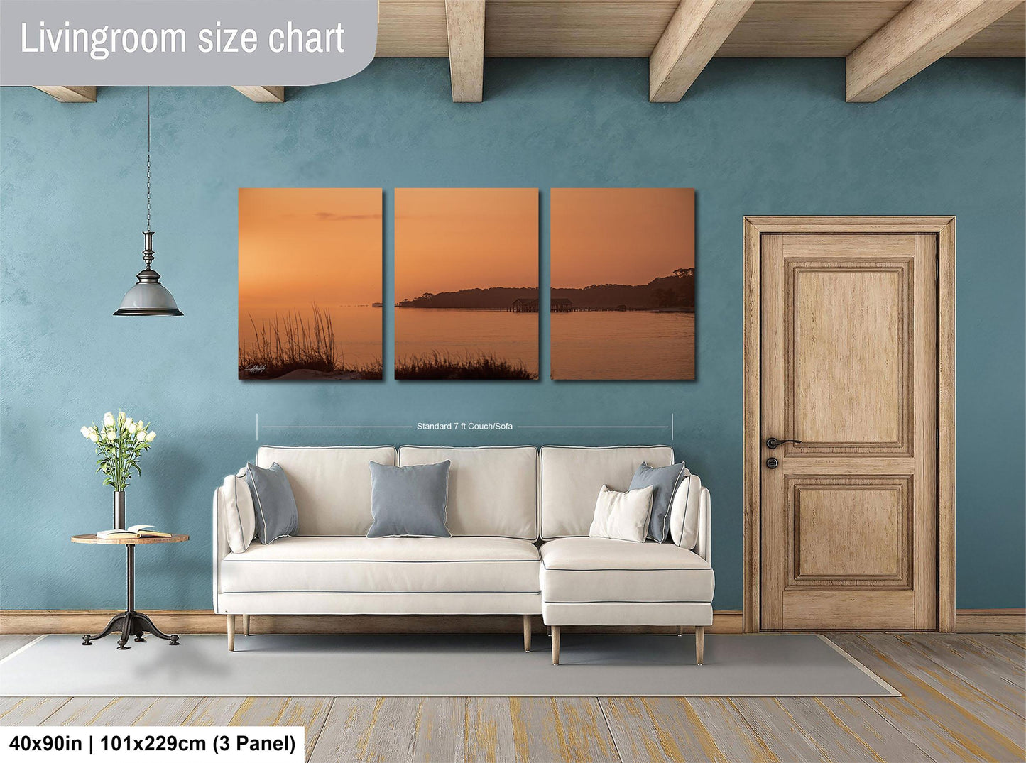 The image is a living room with a large wall art piece depicting a sunset over a body of water. The living room has a white sofa, a wooden door, and a small table with a vase of flowers. The wall art is approximately 40 inches wide and 101.29 cm tall, with a 3-panel layout.