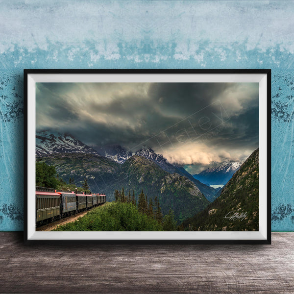 A framed photograph depicting a train traveling through a mountainous landscape with snow-capped peaks and a dramatic cloudy sky.