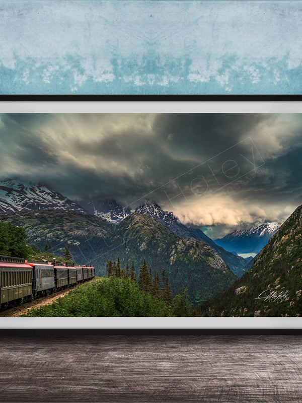 A framed photograph depicting a train traveling through a mountainous landscape with snow-capped peaks and a dramatic cloudy sky.