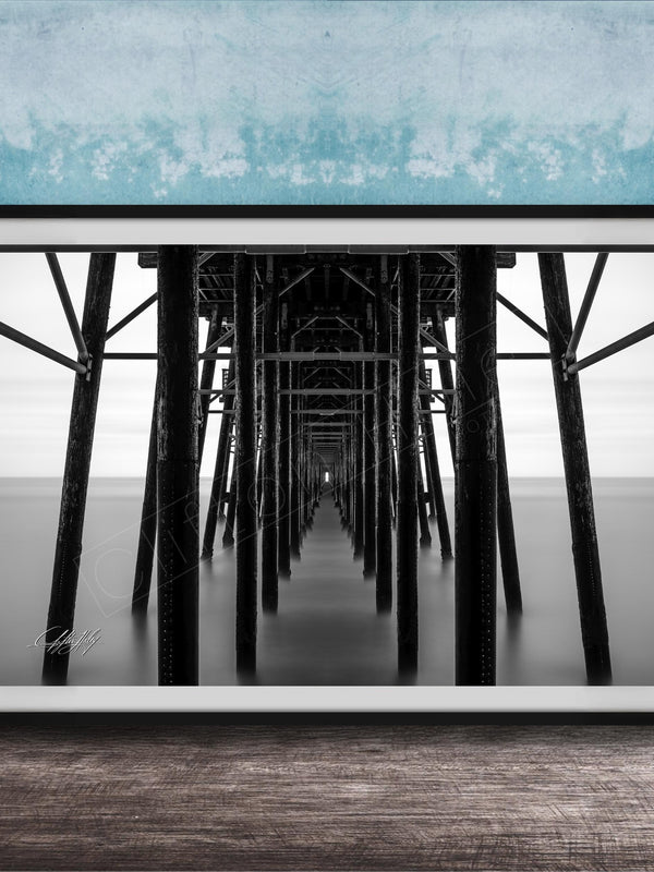 A black and white photograph of a long pier with multiple supporting columns, creating a sense of depth and perspective.