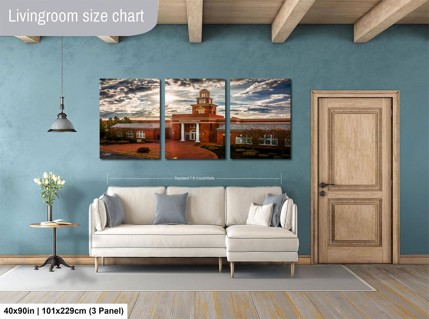 The image is a living room with a large white sofa and a wooden door. Above the sofa, there is a wall-mounted three-panel artwork depicting a building with a clock tower. The artwork is framed in a standard size of 71.25 x 113.29 cm (3 x 3 panel).
