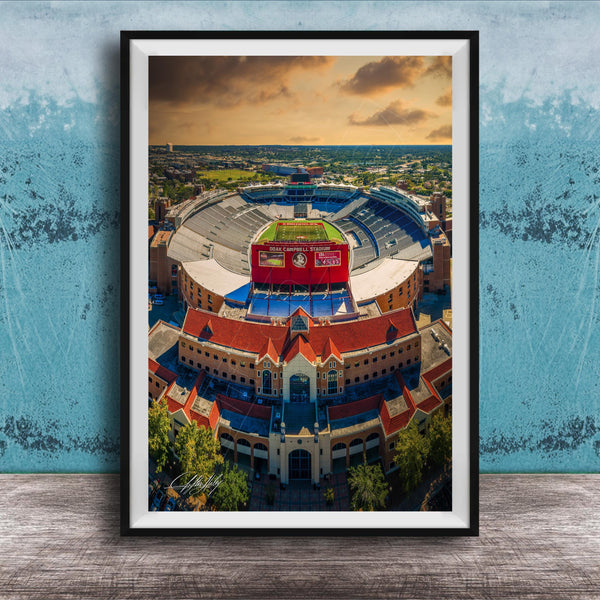 The image is an aerial view of a large stadium with a red roof, surrounded by buildings and a blue sky with clouds.