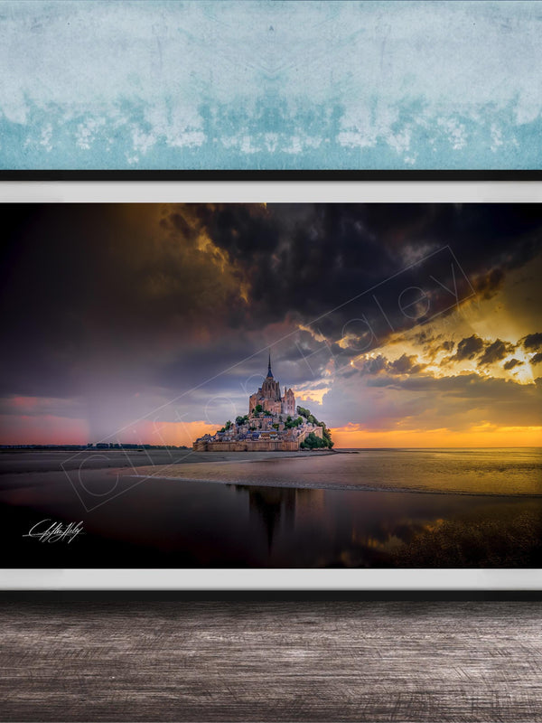 A framed photograph depicting the iconic Mont Saint-Michel island at sunset, with the island's reflection visible in the calm waters below.