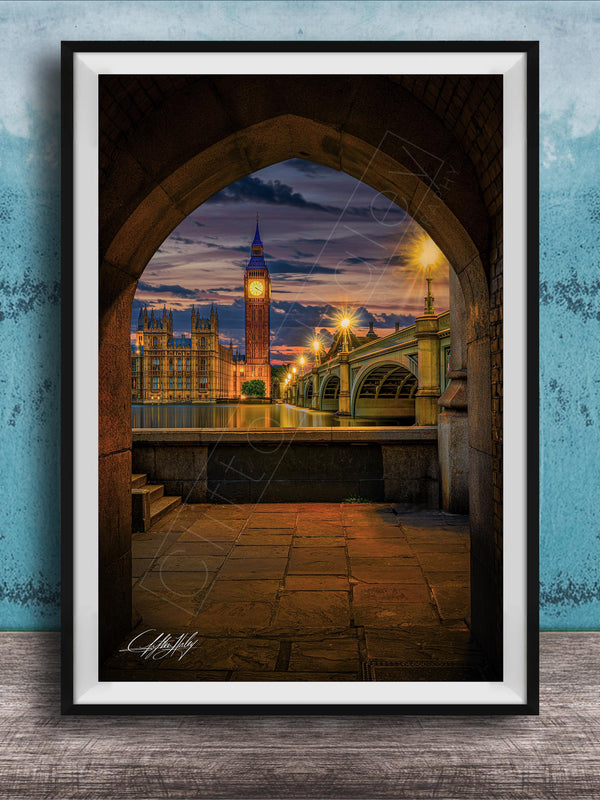 a cityscape at night, with a clock tower and a bridge visible in the background. The clock tower is illuminated, and the bridge is lit up, creating a warm and inviting atmosphere. The scene is framed in a black frame, which contrasts with the blue wall in the background. The image is a collage of multiple images, and the artist's signature is visible in the bottom right corner.