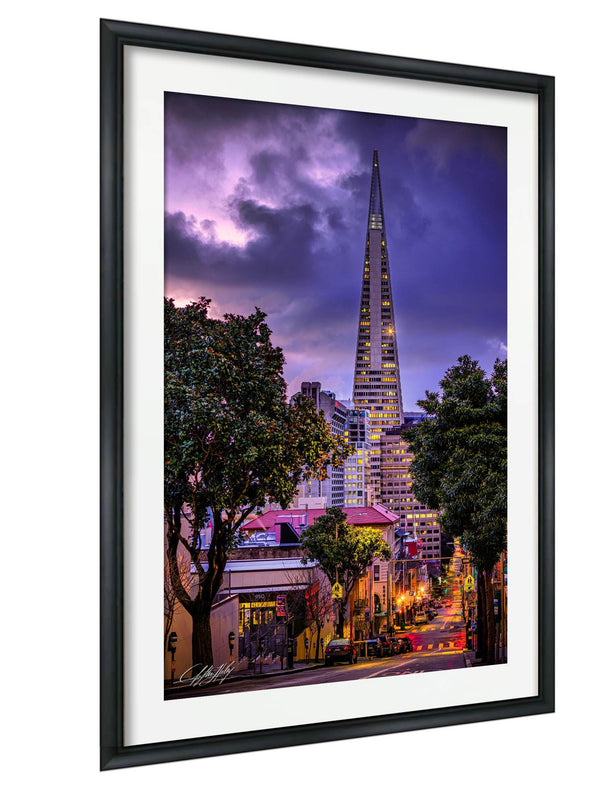 a cityscape at night, with a tall, illuminated skyscraper standing prominently in the background. The city is surrounded by trees and buildings, creating a vibrant and lively atmosphere. The sky above is filled with clouds, adding a dramatic touch to the scene. The image is framed, suggesting it is a piece of art or photography.