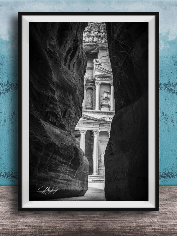 A black and white photograph of a narrow passage between two large rock formations, with a classical building visible through the opening.