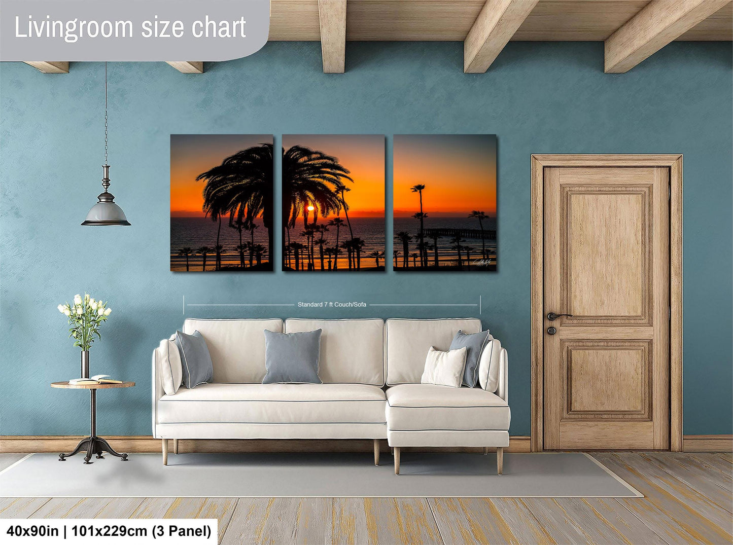 The image is a living room with a white sofa and a wooden door. Above the sofa, there is a large wall art depicting a sunset scene with palm trees silhouetted against the sky. The wall art is composed of three panels, each showing a different part of the sunset. The living room has a blue wall and a wooden floor, creating a cozy and inviting atmosphere.