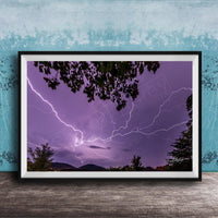 A framed photograph depicting a dramatic lightning storm with vibrant purple and blue hues illuminating the night sky, framed against a blue wall.