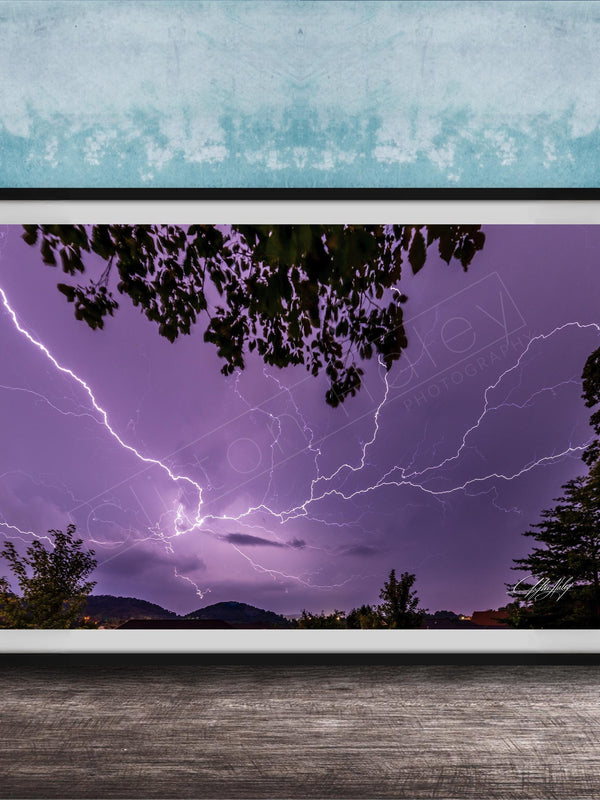A framed photograph depicting a dramatic lightning storm with vibrant purple and blue hues illuminating the night sky, framed against a blue wall.