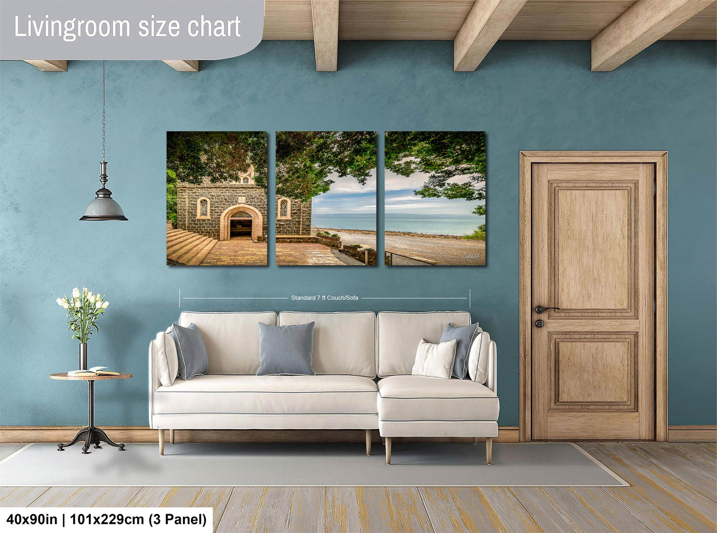 The image is a living room with a white sofa and a wooden door. Above the sofa, there is a triptych of landscape paintings depicting a beach and a building. The wall behind the sofa has a blue color, and there is a lamp hanging from the ceiling. The image is described as a living room size chart, indicating that it is a scale model or a representation of the actual living room size.