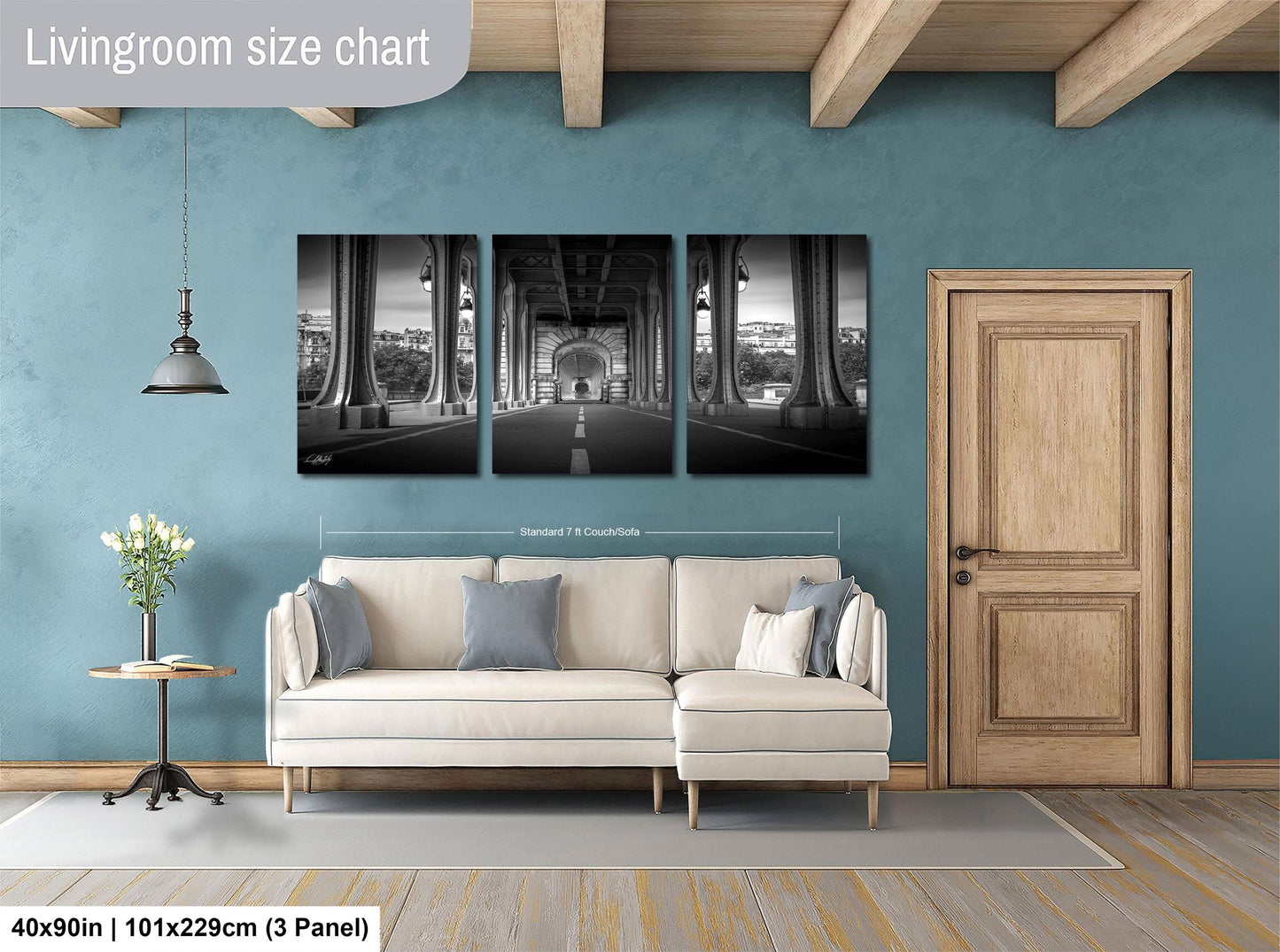 The image is a living room with a large white sofa, a wooden door, and a blue wall. Above the sofa, there is a triptych of black and white photographs depicting a bridge and a building. The living room appears to be well-lit and spacious, with a pendant light hanging from the ceiling and a vase of flowers on a side table.