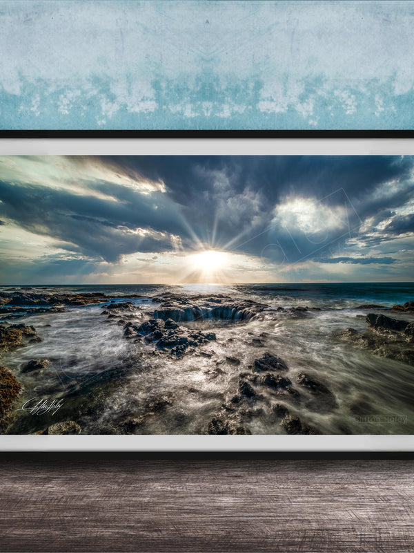 The image is a framed photograph of a rocky coastline with crashing waves, set against a backdrop of a cloudy sky and the sun peeking through the clouds.