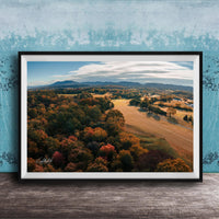 The image is a framed aerial view of a scenic landscape with a mix of trees, fields, and mountains in autumn colors.
