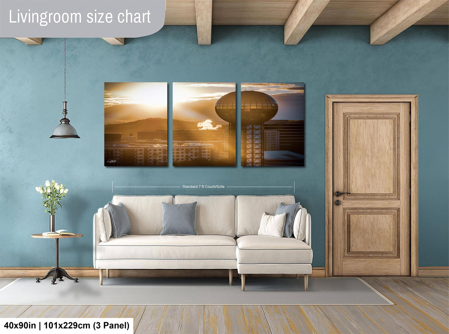 The image is a living room with a large, three-panel wall art depicting a city skyline at sunset. The wall art is mounted above a white sofa, and there is a wooden door and a small table with a vase of flowers nearby. The living room has a blue wall and wooden flooring.