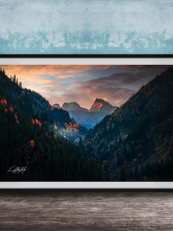 The image is a framed photograph of a mountainous landscape with a sunset sky, featuring a valley with trees and mountains in the background.