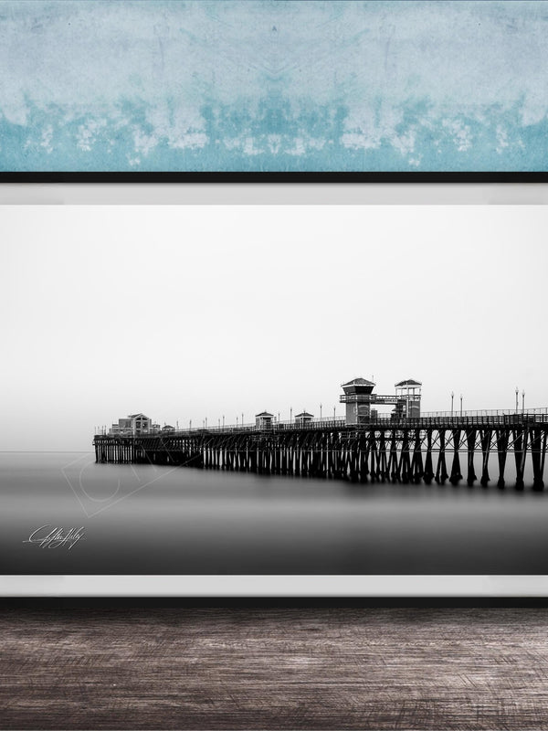 A black and white photograph of a long pier extending into the distance, with a calm body of water below.
