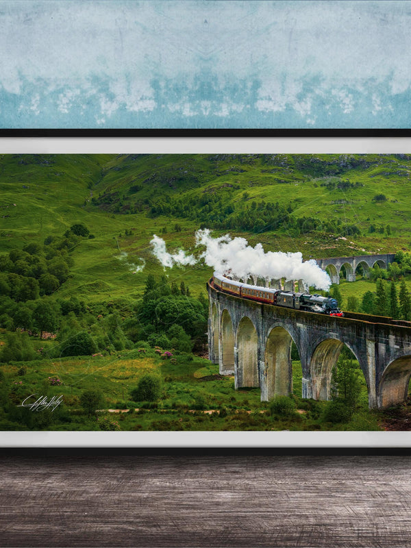 A framed photograph depicting a steam train traveling on a stone bridge over a lush, green valley.