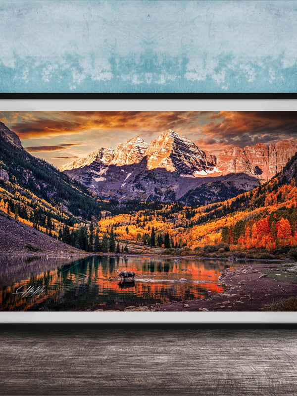 a breathtaking mountain landscape with a lake in the foreground, surrounded by vibrant autumn foliage. The mountains are covered in snow, creating a stunning contrast against the colorful sky. The scene is framed in a black frame and displayed on a wooden surface, adding to the rustic charm of the setting.
