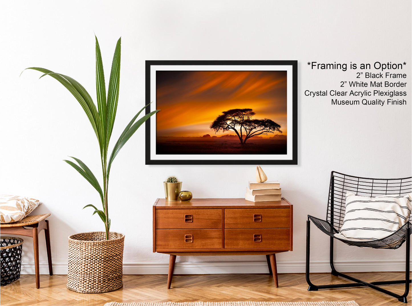 a serene indoor setting with a large framed picture of a tree against a sunset sky hanging on a white wall. The picture is framed in a black frame, and the wall is painted white. In front of the picture, there is a wooden dresser with a wicker basket on top. To the right of the dresser, there is a black wire chair with a striped pillow on it. A potted plant is also present in the scene.