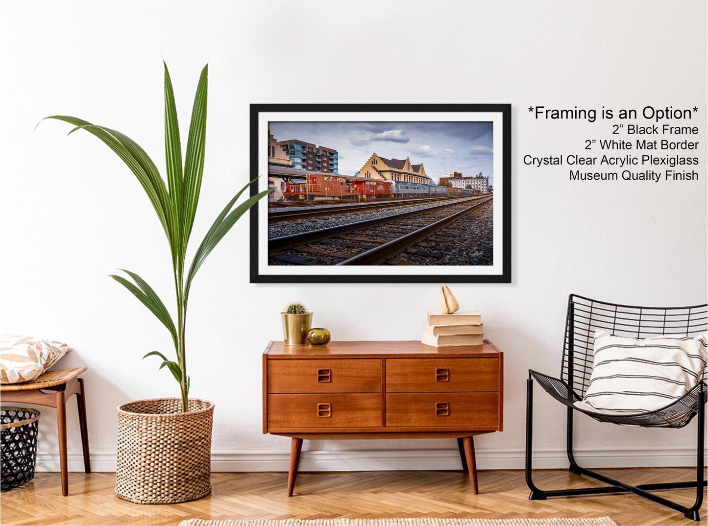 a framed photograph of a train station with a black frame, a white border, and a crystal clear acrylic plexiglass finish. The photograph is hung on a white wall above a wooden dresser with a wicker basket. A black wire chair is positioned next to the dresser, and a potted plant is placed on the floor next to the chair.